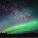 ホワイトホースの夜空に広がる緑のオーロラと星空、森のシルエットが映える冬の風景