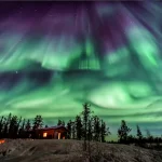 夜空に広がるオーロラの幻想的な風景。森と小屋の上に揺らめく緑の光が広がり、冬の絶景を楽しめる人気の自然現象。
