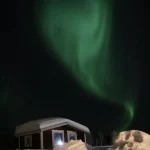 雪に包まれたキャビンの上空に緑のオーロラが広がる冬の夜景
