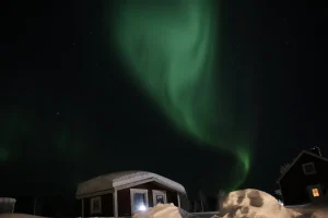 雪に包まれたキャビンの上空に緑のオーロラが広がる冬の夜景
