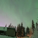 雪に覆われた森とロッジの上空に淡いオーロラが広がる冬景色