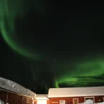 雪に包まれたロッジの上空に緑のオーロラが広がる冬の夜景
