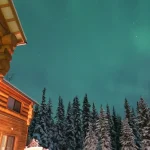 雪に包まれたロッジと森の上にオーロラが広がる冬の旅行風景