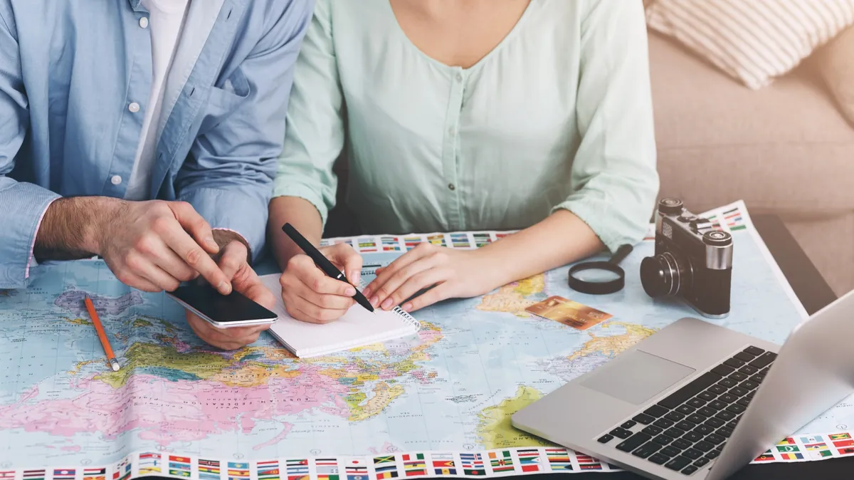 世界地図やPCを広げて旅行日程を相談するカップルのハネムーン計画イメージ