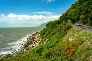ケアンズ近郊の海岸沿いを走る車と青い海、緑豊かな山並みが広がるドライブ風景