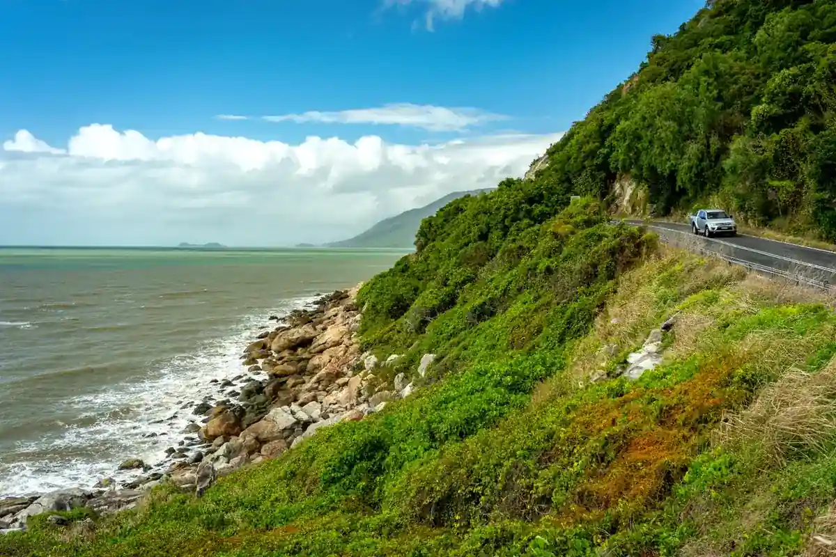 ケアンズ近郊の海岸沿いを走る車と青い海、緑豊かな山並みが広がるドライブ風景