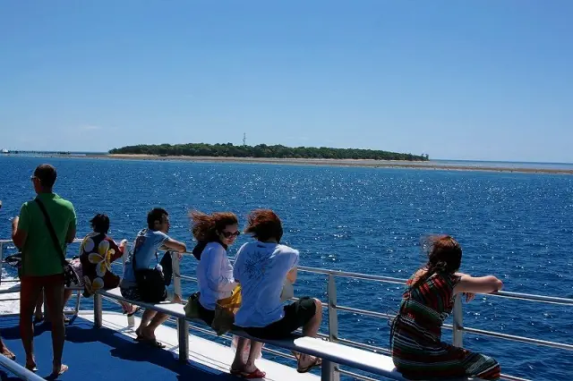 船の屋外デッキから青い海と水平線近くに広がる島を眺める乗客たち